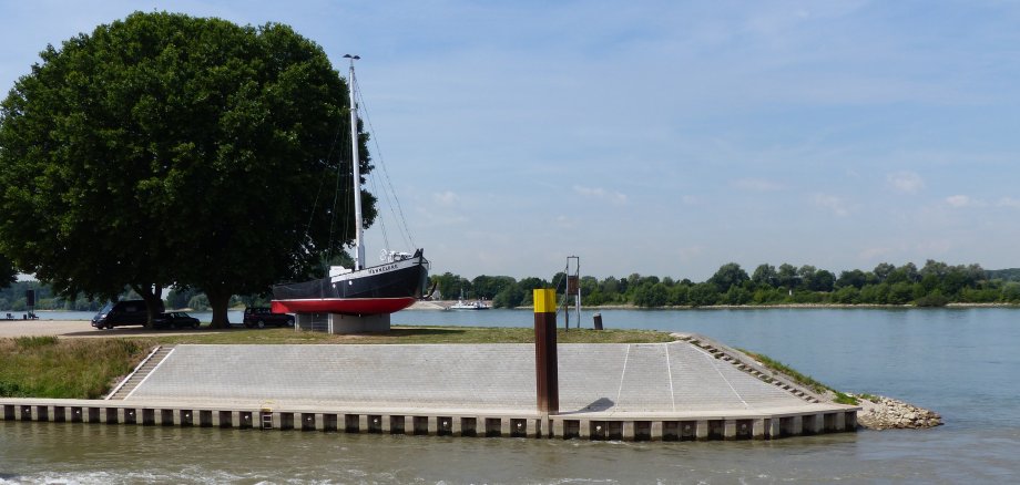 In der Mitte befindet sich die Hafenspitze von Gernsheim umgeben von Wasser des Rheins. In der Mitte der Hafenspitze befindet sich auf einem Betonsockel ein altes Fischerboot mit Namen Hannelore. Links davon stehen zwei große Bäume unter denen drei Autos parken. 